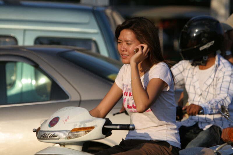 Talking while Driving in Cambodia — Woman talks on her phone while driving her motor scooter in Phnom Penh, Cambodia — Cambodia, street, streets, traffic, mo...