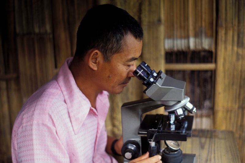 Man at Microscope in Thailand — Stock Image of a health worker at an remote ADRA Clinic set up in a bamboo shelter at a Karen Refugee camp, checks microscope...