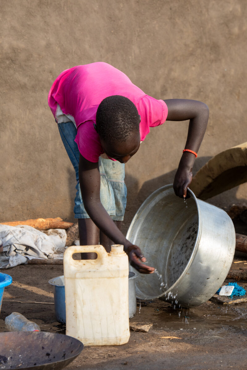 Washing Dishes — Young refugee girl washes dishes in her new home in Uganda. — Person, Refugee, Refugee Settlement, South Sudan