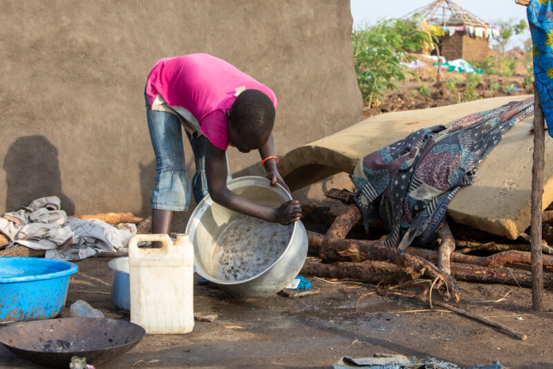 Washing Dishes — Young refugee girl washes dishes in her new home in Uganda. — Animal, Person, Reptile, Refugee, Refugee Settlement