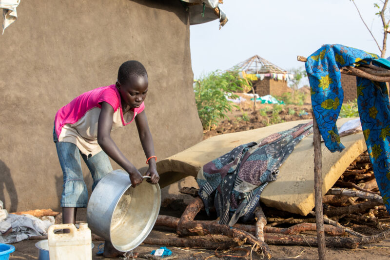 Washing Dishes — Young refugee girl washes dishes in her new home in Uganda. — Eyes Closed, Female, Frontal Face, One Face, Person