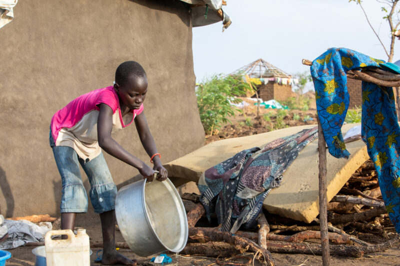 Washing Dishes — Young refugee girl washes dishes in her new home in Uganda. — Animal, Child, Eyes Closed, Female, Frontal Face