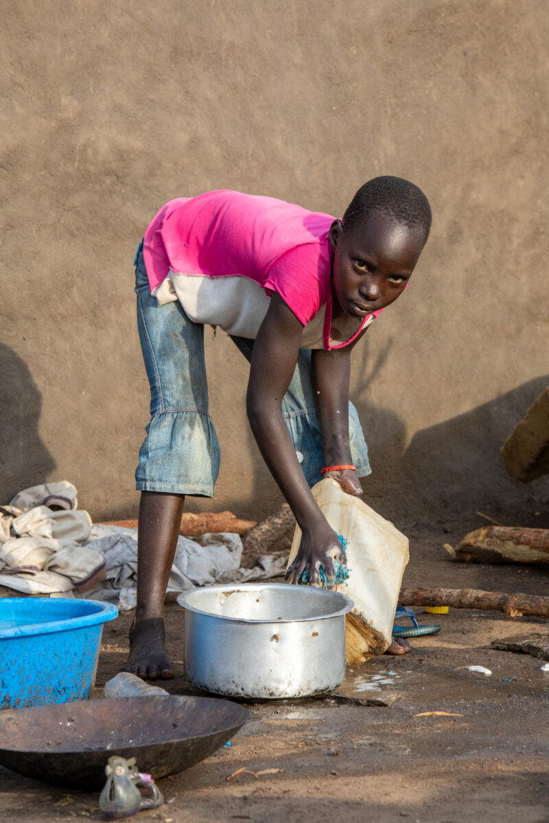 Washing Dishes — Young refugee girl washes dishes in her new home in Uganda. — Child, Eyes Open, Female, Frontal Face, One Face