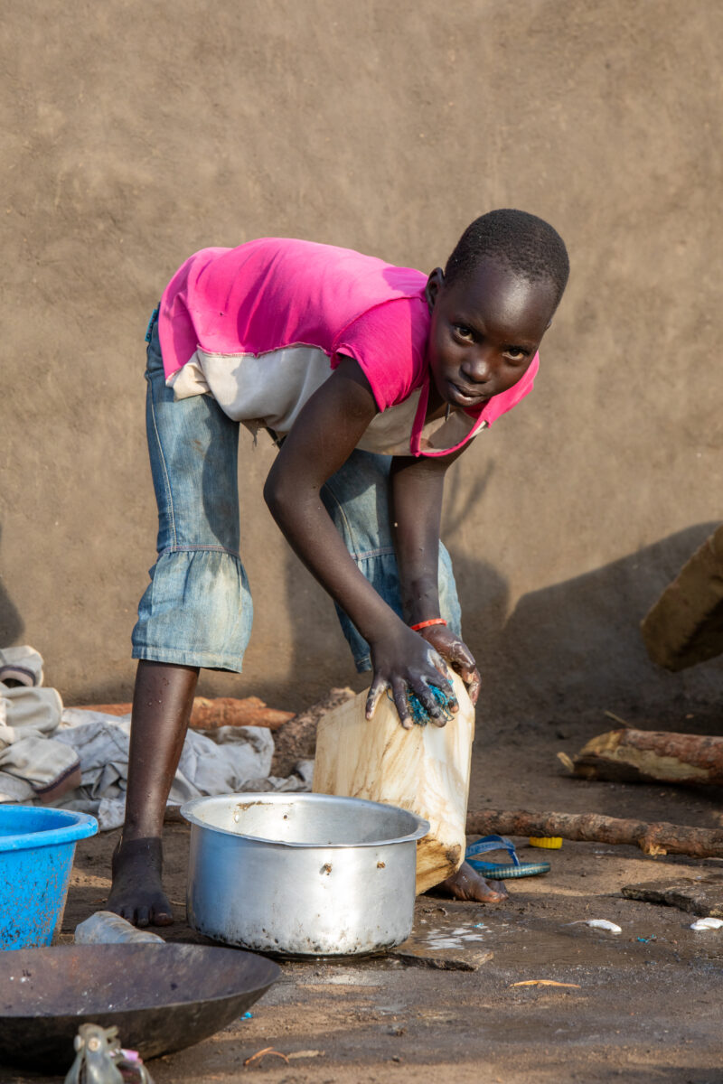 Washing Dishes — Young refugee girl washes dishes in her new home in Uganda. — Eyes Open, Female, Frontal Face, One Face, Person