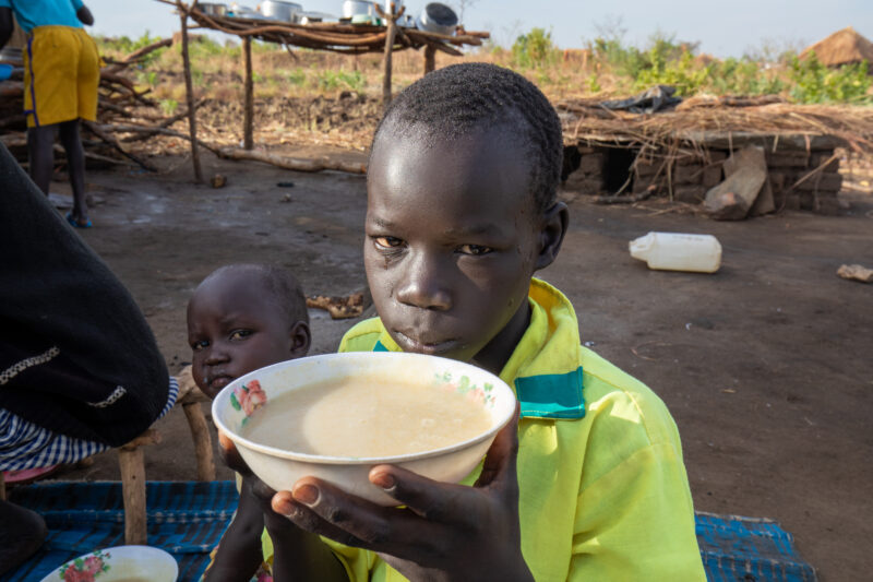 Refugee Child Eating — Refugee from South Sudan takes a morning meal of porridge — Adult, Child, Eyes Open, Food, Frontal Face