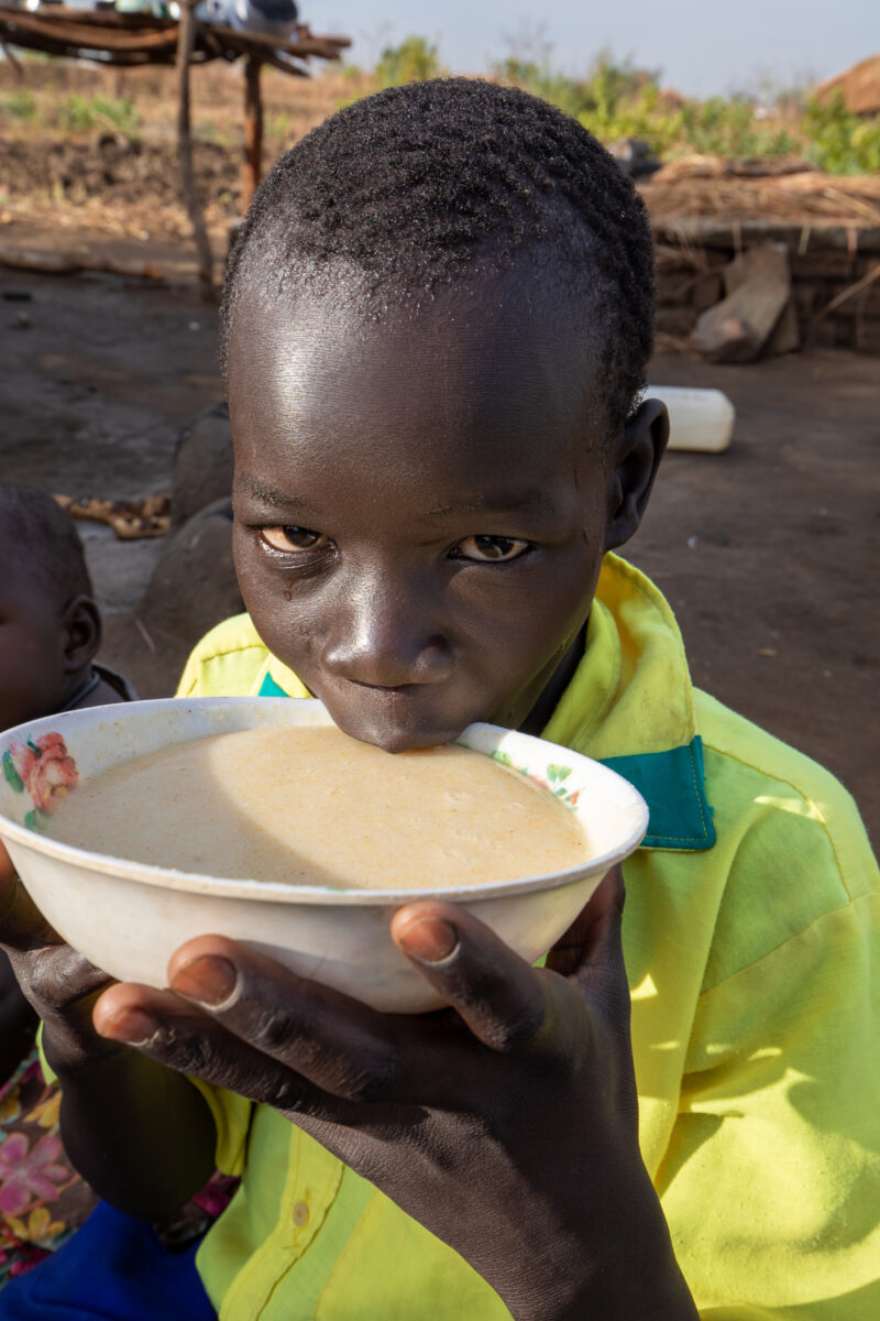 Refugee Child Eating — Refugee from South Sudan takes a morning meal of porridge — Female, Food, Frontal Face, One Face, Person
