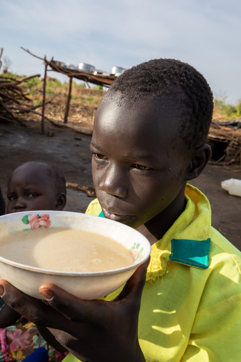 Refugee Child Eating — Refugee from South Sudan takes a morning meal of porridge — Child, Eyes Open, Female, Food, Frontal Face
