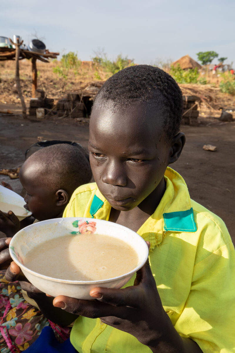 Refugee Child Eating — Refugee from South Sudan takes a morning meal of porridge — Adult, Eyes Open, Food, Frontal Face, Male