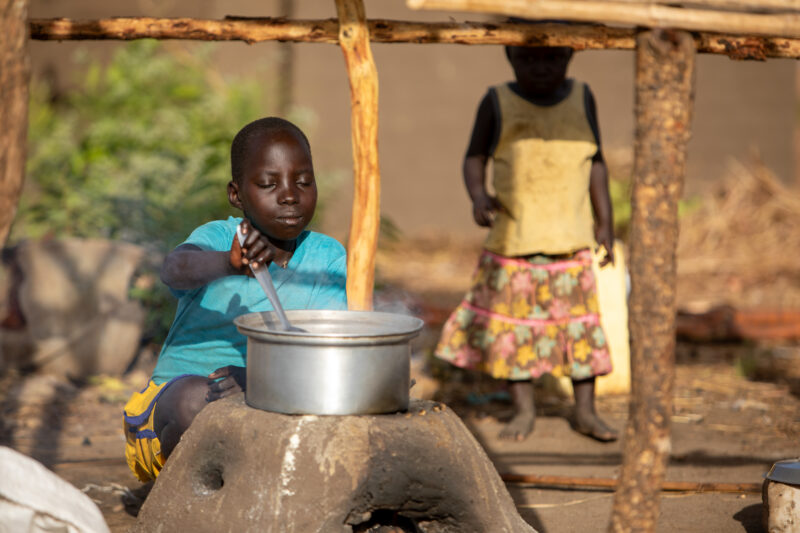 Cooking the Meal — Young refugee girl cooks a meal outdoors on a mud based cooking stove. — Child, Eyes Closed, Frontal Face, Male, One Face