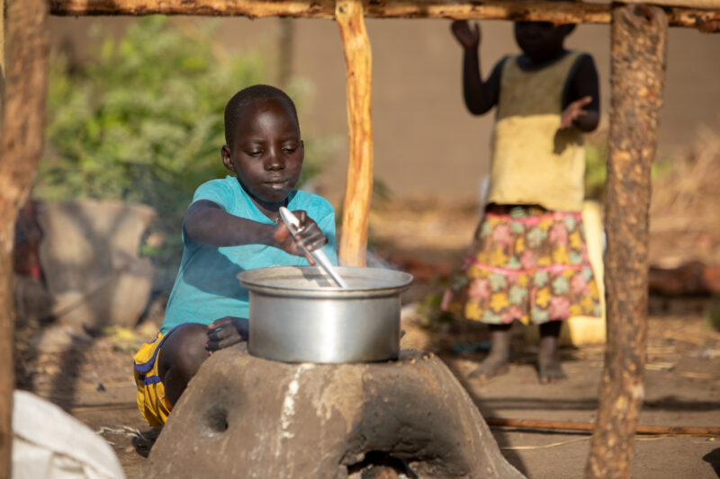 Cooking the Meal — Young refugee girl cooks a meal outdoors on a mud based cooking stove. — Child, Eyes Closed, Frontal Face, Male, One Face
