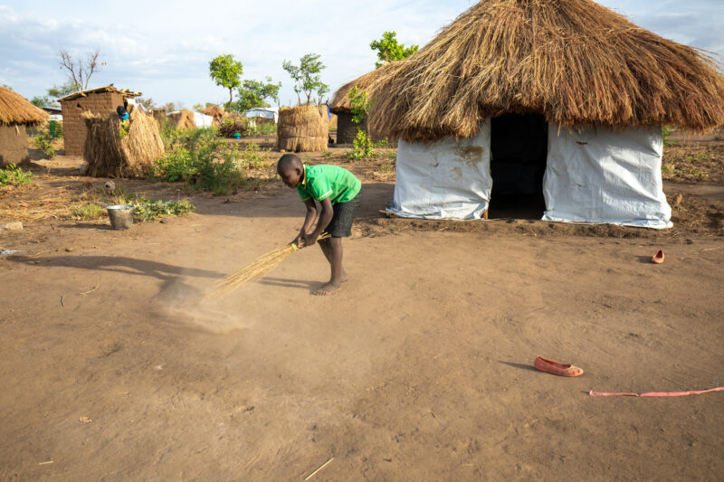 Sweeping the Yard — Young Refugee girl sweeps the yard outside the small house they have built in a refugee settlement in Uganda. — Person, Refugee, Refugee ...