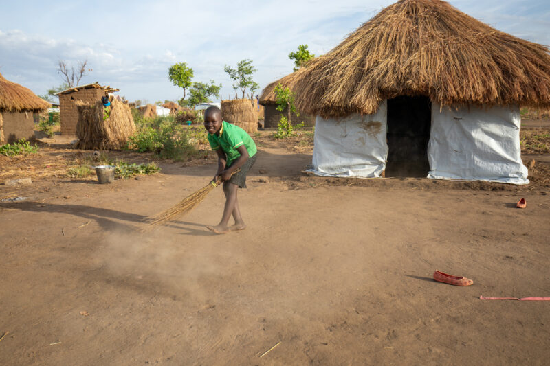 Sweeping the Yard — Young Refugee girl sweeps the yard outside the small house they have built in a refugee settlement in Uganda. — Child, Eyes Open, Female,...