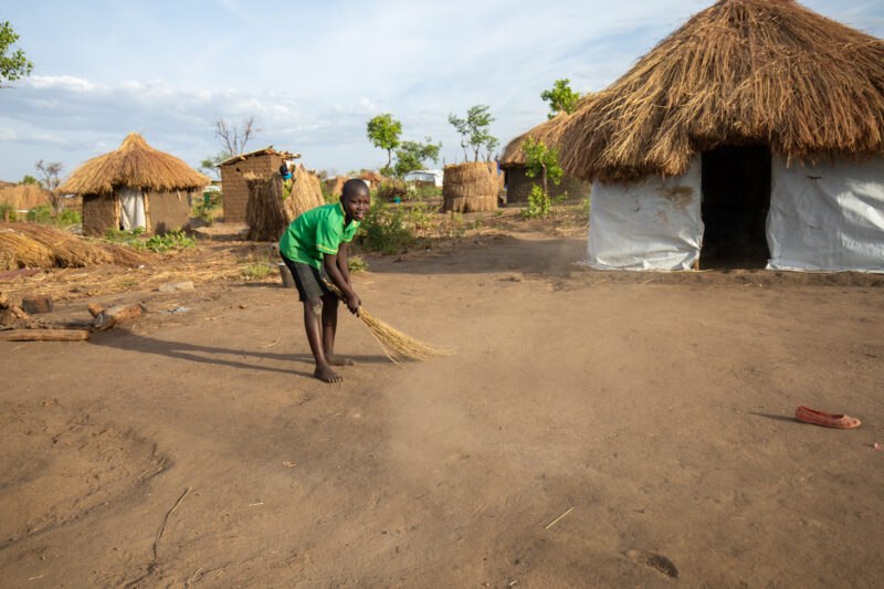 Sweeping the Yard — Young Refugee girl sweeps the yard outside the small house they have built in a refugee settlement in Uganda. — Eyes Open, Female, Fronta...