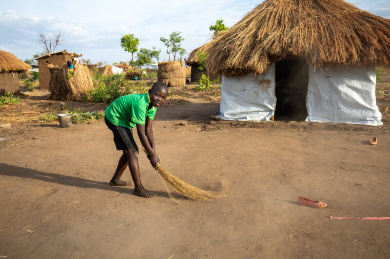Sweeping the Yard — Young Refugee girl sweeps the yard outside the small house they have built in a refugee settlement in Uganda. — Child, Eyes Open, Female,...