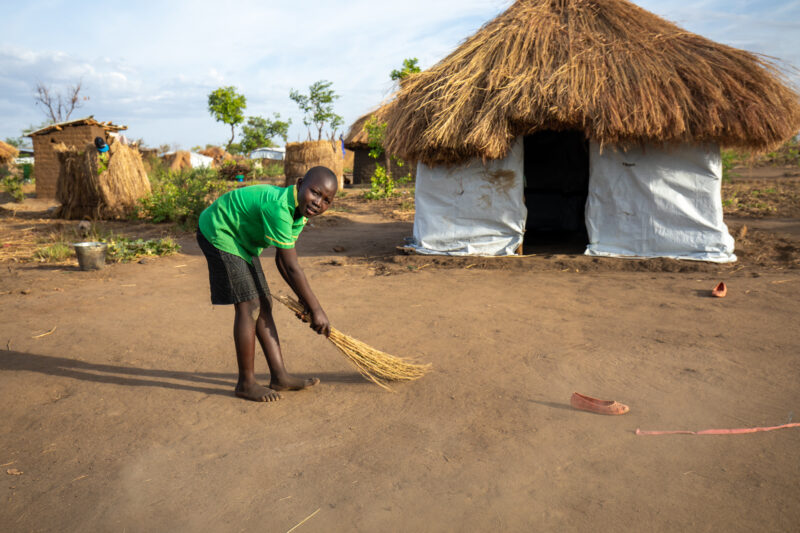 Sweeping the Yard — Young Refugee girl sweeps the yard outside the small house they have built in a refugee settlement in Uganda. — Eyes Open, Female, Fronta...