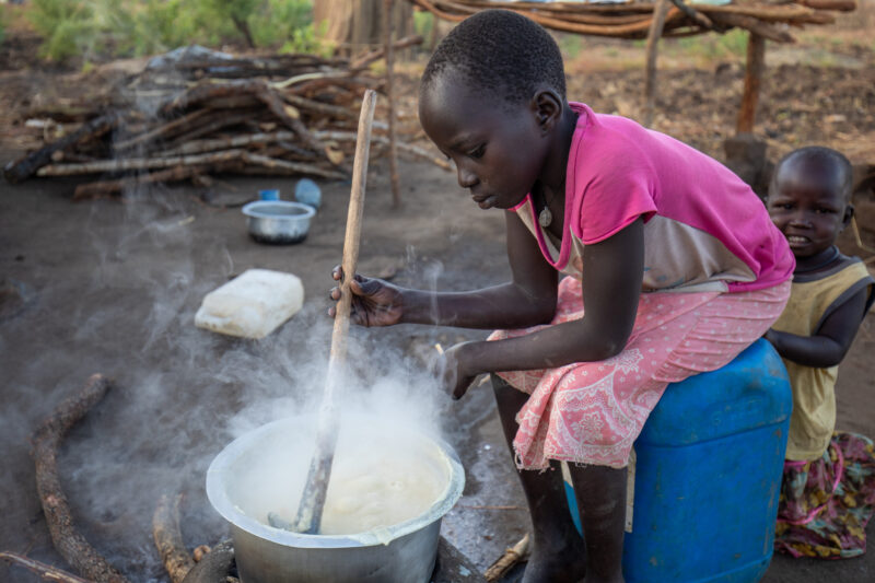 Cooking the Meal — Young refugee girl cooks a meal outdoors on a mud based cooking stove. — Child, Eyes Open, Food, Frontal Face, Male