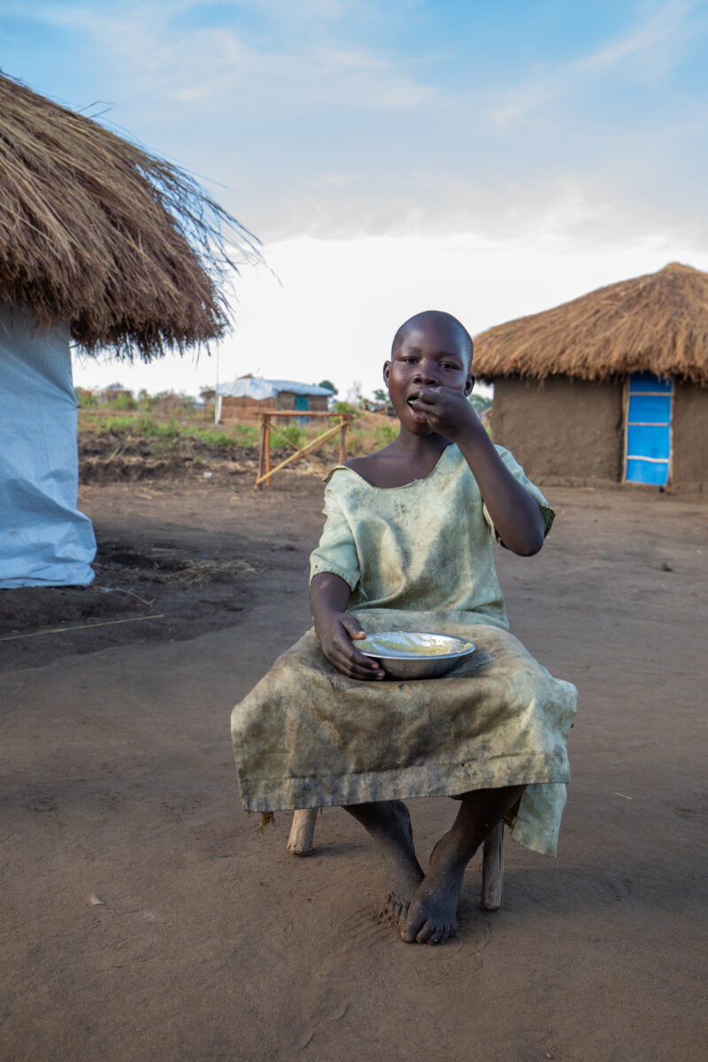 Refugee Child Eating — Refugee from South Sudan takes a morning meal of porridge — Eyes Open, Frontal Face, Kneeling, Male, One Face