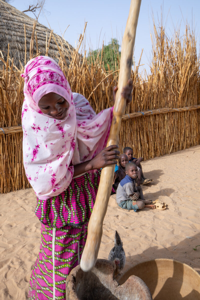 Pounding the Meal — Girl pounds sorgum into powder with a pounding stick — Africa, Child, Education, Eyes Closed, Eyes Open