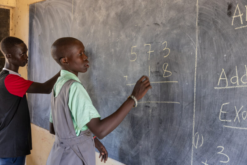 Doing Math on the Board — Young girl in South Sudan does a math problem on the blackboard of her school. — Person, Profile Face, Seminar Room, South Sudan, T...