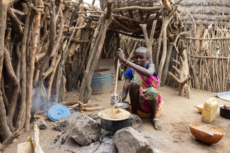 Cooking Breakfast in Rural Africa — Young girl cooks sorghum breakfast over an open fire in the compound of her home. — Frontal Face, One Face, Person, South...