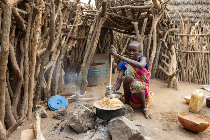Cooking Breakfast in Rural Africa — Young girl cooks sorghum breakfast over an open fire in the compound of her home. — Frontal Face, One Face, Person, South...