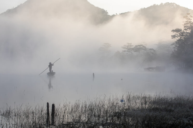 Early Morning on the Lake — Fog and mist rise up from a Lake in Vietnam making a scenic splender with a man in his fishing boat. — Fog, Nature, Vietnam, Scenic