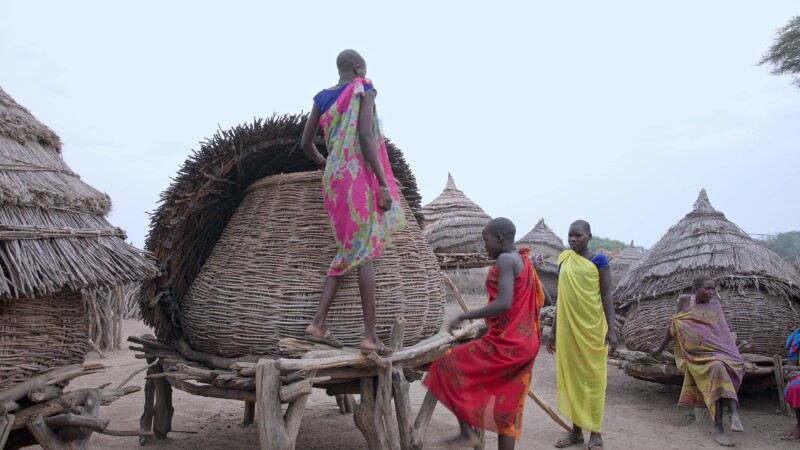 Grainery — Young girls climbs into a food storage hut to get grain for cooking. — Africa, South Sudan, Rural, Remote, Pastoralists